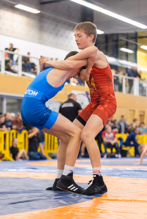 Two youth wrestlers locked in an intense clinch, red vs blue singlets, competing on an orange mat before a crowd.