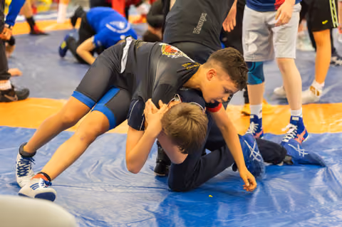 Two young wrestlers grapple on a blue mat, one boy dominating with focused intensity as he pins his opponent down.