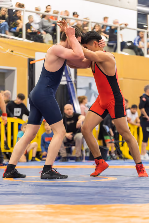 Two wrestlers in navy and red singlets lock arms in an intense clinch on a blue competition mat, crowd watching.