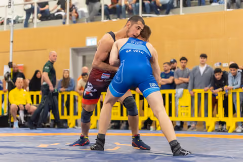 Two wrestlers lock up intensely on the mat; blue SWE BK Velo athlete engages a red-singlet opponent before a crowd.