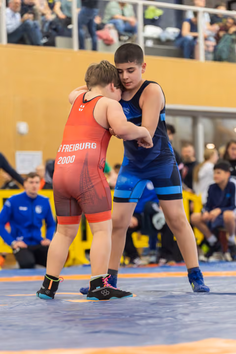 Two youth wrestlers engage in a standing clinch on the mat, one in red Freiburg 2000 singlet, the other in blue.