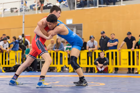 Two young wrestlers locked in a tight clinch on the mat, both leaning forward with intense focus during a competitive bout.
