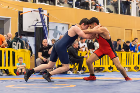 Two wrestlers clash forehead-to-forehead in intense clinch, one in navy, one in red, on a blue mat before a crowd.