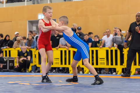 Two young boys lock wrists in an intense standup grapple on a blue mat, watched by a crowded audience.