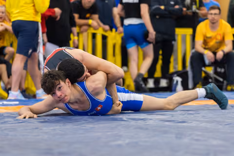 Young Swiss wrestler in blue singlet pinned on the mat, straining against opponent, crowd watching in background.