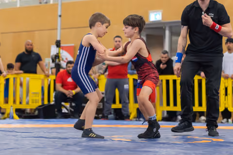 Two focused young boys in singlets grip each other's arms on a blue mat, referee watching closely nearby.