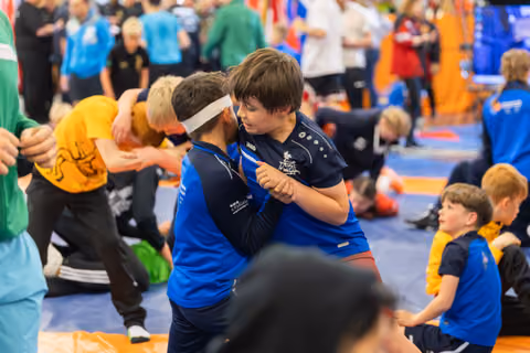 Two young boys in blue Jako jerseys lock grips, faces close, intense concentration during a busy indoor wrestling event.