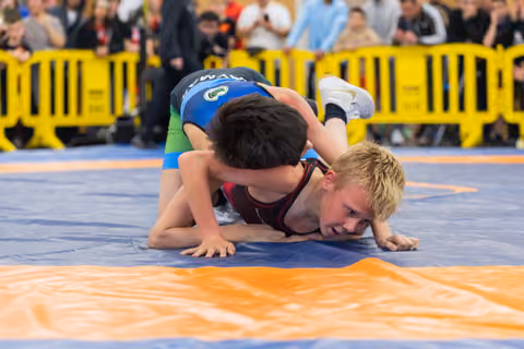 Two young boys wrestling on a blue mat, one pinning the other who strains with effort and determination.