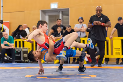Wrestler in red lifts opponent in blue off the mat in a powerful throw, referees and crowd watching intently.