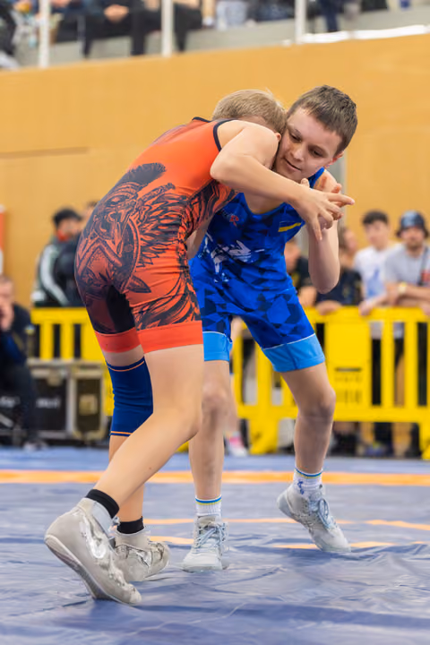 Two young wrestlers locked in a tight clinch on the mat, one in red, one in blue, both straining with focused determination.