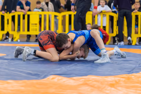 Two young boys in red and blue singlets locked in an intense grapple on a blue mat, crowds watching behind barriers.
