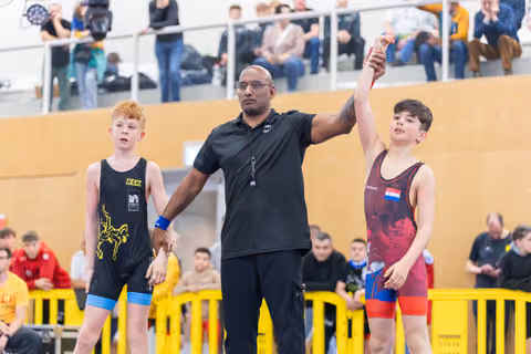 Referee raises the arm of a victorious boy in red, while the runner-up in black stands beside him, indoors.