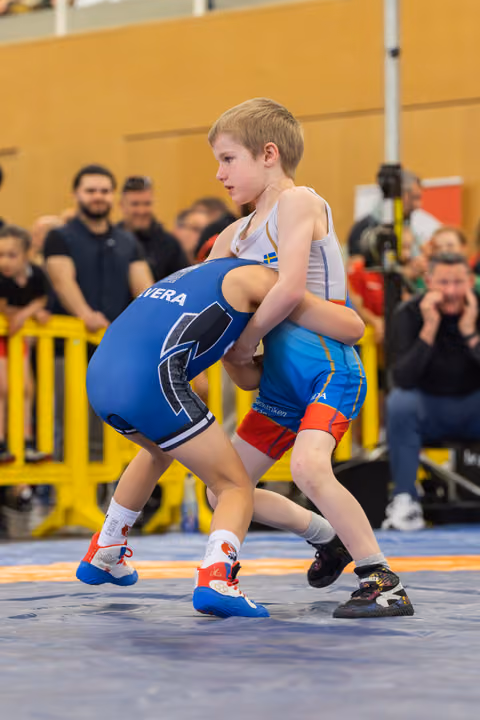 Two young boys locked in an intense wrestling grip on a blue mat, spectators watching closely in the background.