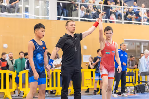Referee raises the arm of a young wrestler in red, declaring victory, while the opponent in blue looks on stoically.