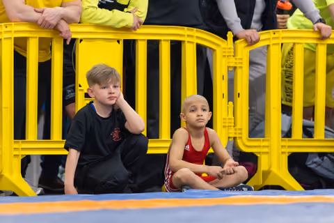 Two young boys sit cross-legged on the mat beside yellow barriers, watching the event with focused, serious expressions.