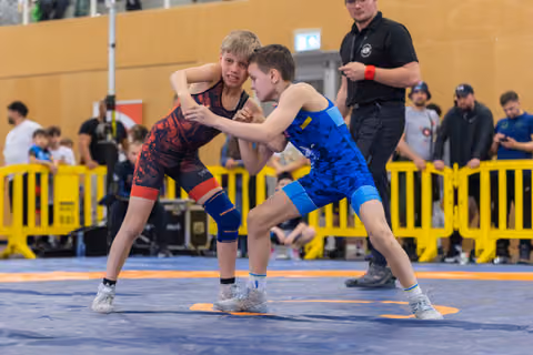 Two young wrestlers in red and blue singlets lock grips on the mat, focused and determined during a youth bout.