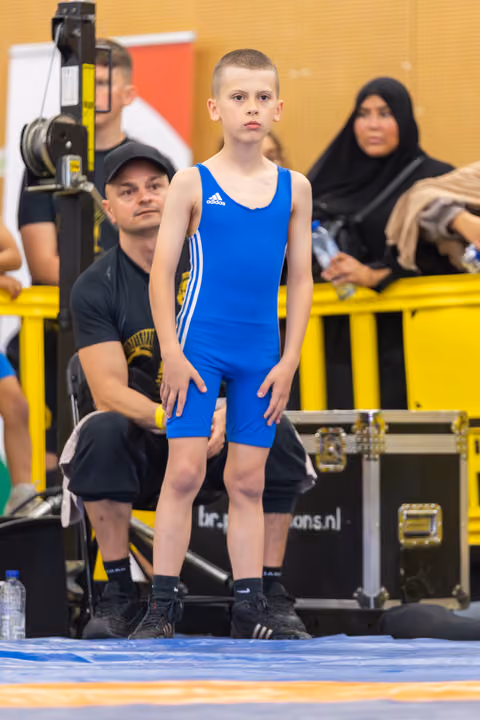 Young boy in blue Adidas singlet stands poised on the mat, composed and focused, coach crouching attentively behind him.