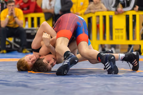 Two young wrestlers grapple on the mat, one dominant on top controlling the other in a tight hold, both straining with effort.