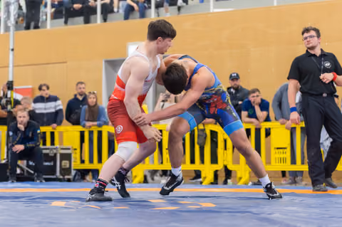 Two young wrestlers locked in intense grappling, one in red singlet, one in blue, battling on a blue competition mat.