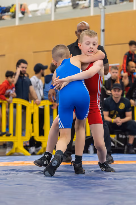 Two young boys locked in a grappling clinch, focused and determined, wrestling on a blue mat before a crowd.