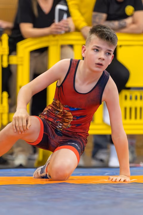 Focused young boy in red singlet kneels on the mat, alert and ready, with spectators seated behind yellow barriers.