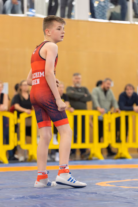 Young boy in red singlet stands alert on the mat, fists clenched, focused and ready during a youth wrestling event.