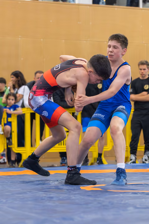 Two teenage wrestlers grapple intensely on a blue mat, one in black-red, the other in blue, spectators watching behind barriers.
