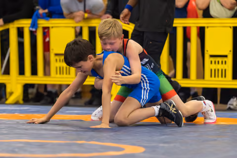 Two young boys wrestling on a blue mat, one boy in blue singlet struggles forward while opponent grips him from behind.