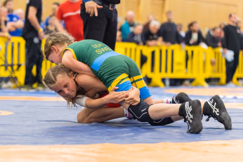 Young girl in Sweden green singlet pins opponent to blue mat, both showing intense focus during youth bout.