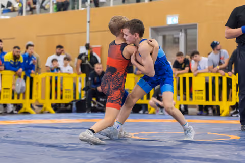 Two young boys locked in a fierce wrestling clinch on a blue mat, crowds watching behind yellow barriers.