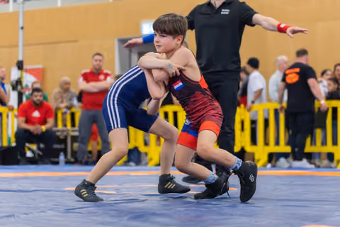 Two young wrestlers lock grips on the mat, focused and determined, as a referee signals in the background.