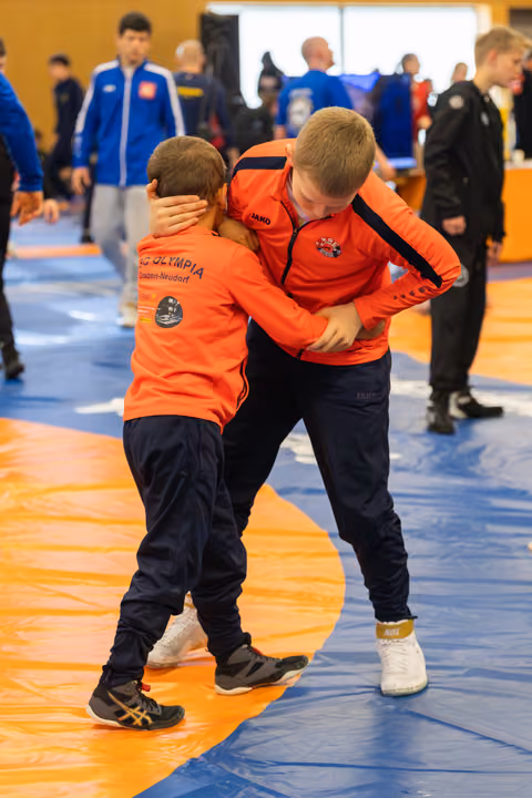 Two boys in orange AC Olympia jackets clinch during a wrestling bout on orange and blue mats, focused and intense.