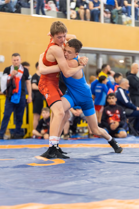 Two teenage wrestlers locked in a tight clinch, straining against each other with intense focus on a blue competition mat.