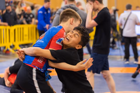 Two young wrestlers grapple intensely on the mat; the boy in black grimaces under strain during an indoor sports event.