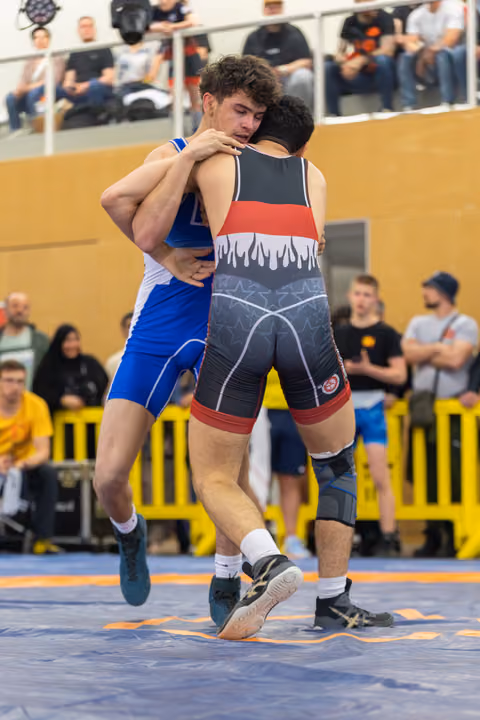 Two wrestlers locked in a tight clinch, one lifted off the mat mid-match, crowd watching in the background.