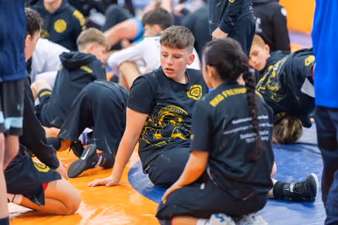 Young wrestlers in black and gold team shirts sit on the mat, chatting between matches at an indoor sports event.
