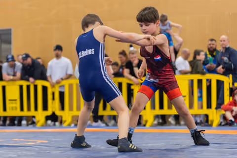 Two young wrestlers engage on the mat, one in blue 'Reims' singlet, the other in red Dutch colors, focused intensity.