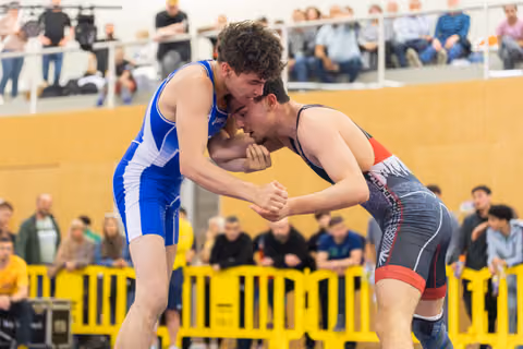 Two young wrestlers lock heads in an intense clinch, both straining with determination on an indoor mat before a crowd.