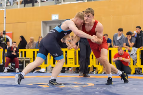 Two male wrestlers in red and blue singlets locked in an intense clinch, both leaning forward with fierce focus on the mat.