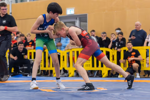 Two young wrestlers face off on a blue mat, one in blue-green, one in red-black, both low and tense.