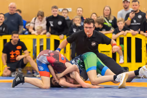 Two young wrestlers grapple intensely on a blue mat, watched closely by a referee and a crowd of spectators.