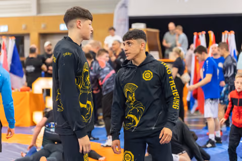 Two teenage wrestlers in black and gold Freiburg tracksuits face each other with focused, determined expressions on the mat.