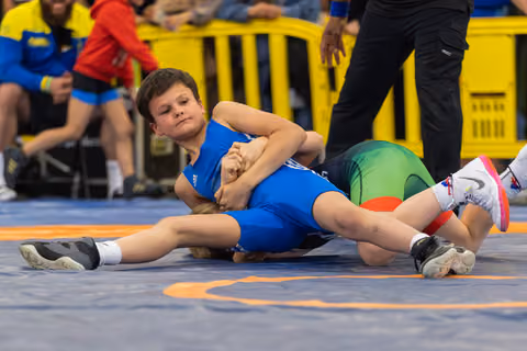 Focused young wrestler in blue singlet pins his opponent on the mat during a youth wrestling match.