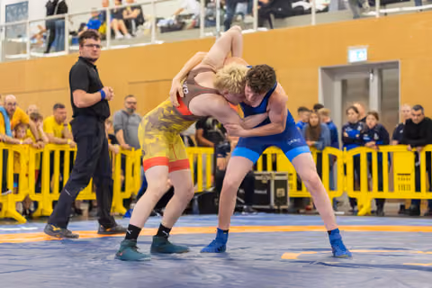 Two young wrestlers locked in a fierce clinch on a blue mat, referee watching closely as crowd looks on.