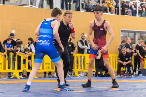 Referee gives instructions to two teen wrestlers—one in blue, one in dark red—before their bout on a blue mat.