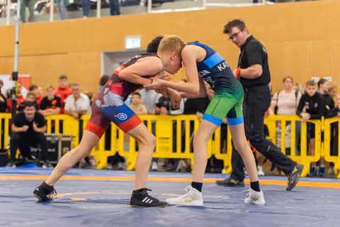 Two young wrestlers lock grips in intense focus on a blue competition mat, referee watching closely behind.