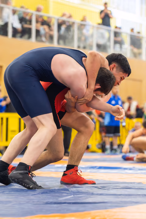 Two wrestlers locked in a tight clinch on the mat, one in navy blue singlet dominating over opponent in red, crowd watching.