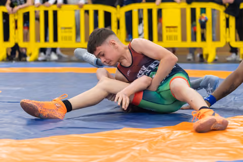 Determined young wrestler controls opponent on blue mat, wearing vibrant orange shoes and red-green singlet.