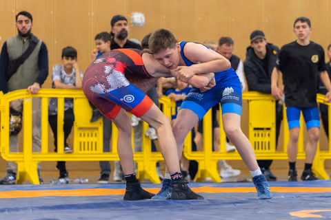 Two young wrestlers in a fierce clinch on the mat, spectators watching behind yellow barriers in a sports hall.