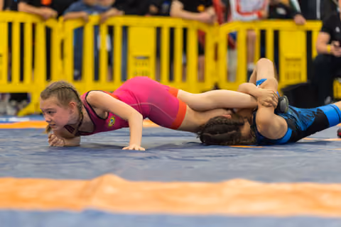 Young girl in pink singlet fights to escape a pin on the blue mat, face showing intense concentration and effort.